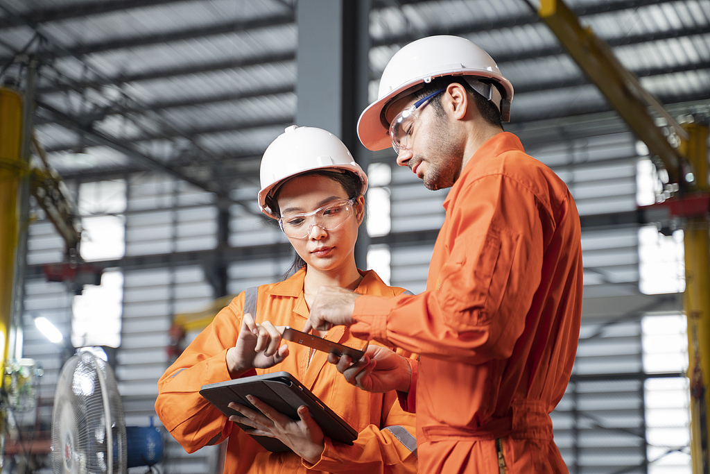 Woman engineer and mechanical technical in overalls using digital tablet and discuss to each other during their working in the factory.